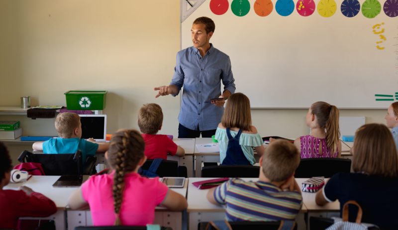 Front view of a Caucasian male school teacher standing and addressing a diverse group of school children sitting at desks during a lesson in an elementary school classroom